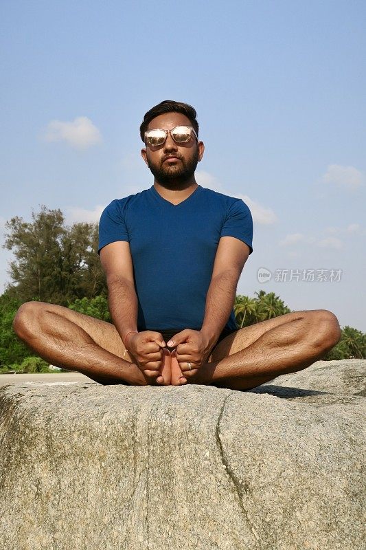 印度男子在岩石上练习蝴蝶/鞋匠姿势(Baddha Konasana)瑜伽姿势，水边，Palolem海滩，果阿，印度